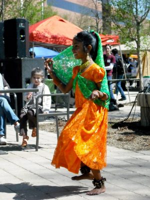 A young girl in bright orange and green clothing from her native country dancing at the street fair.