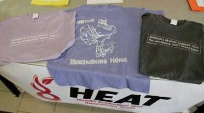 Three T-shirts on a table, one lavendar, which reads, "Surgeon's General Warning: Don't Run with Scissors. Don't Smoke Either.", a purple shirt with the HokieBird stoping a cigarette, and a dark gray shirt that reads, "Surgeon's General Warning: Don't Play with Matches. Don't Smoke Either." 