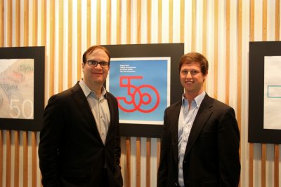 Rocco Piscatello and David Spradlin stand in front of a wall displaying a 50th anniversary icon design. 