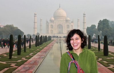 A young woman stands in front of the Taj Mahal in the distance. 