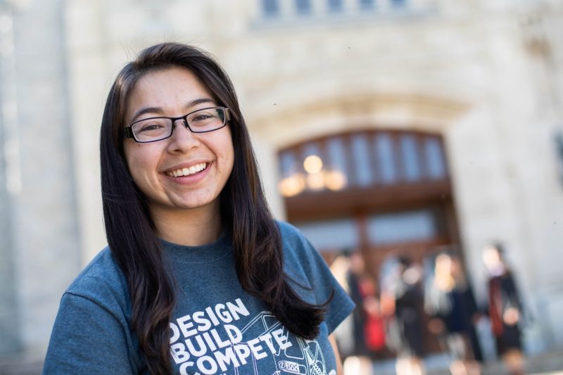 Photo of a smiling young woman standing in front of a gray Hokie stone building, Burruss Hall.