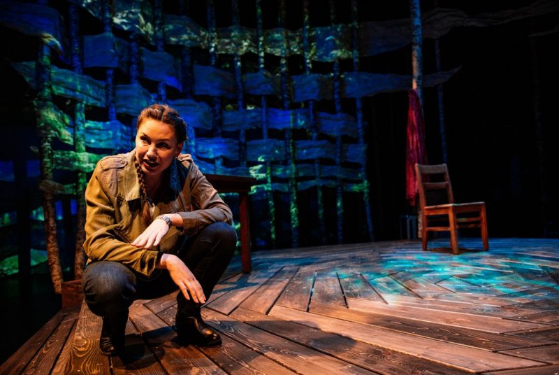 DeLanna Studi crouches on a wooden stage, a lone wooden wooden chair behind her, along with blue- and green-lit backdrop.