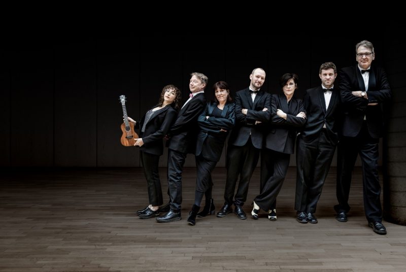 Members of the Ukulele Orchestra of Great Britain, three white women and four white men, stand in a line wearing all black. The women are order from shortest to tallest, left to right, and the men are ordered the same way., and they all lean right towards the tallest man. The woman on the far left holds a ukulele.