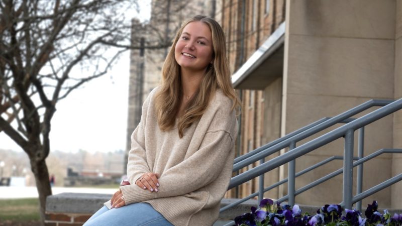 A blond college-aged woman, wearing a beige sweater sits on steps. Purple flowers are in the background.
