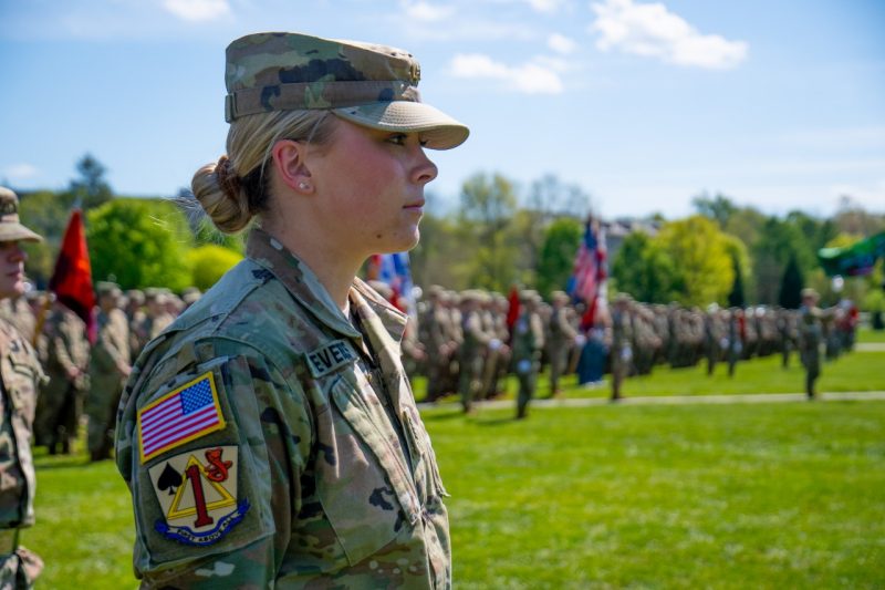 Stevens stands in her camouflage uniform in front of a large group of cadets on the green grass of the Drillfield. 