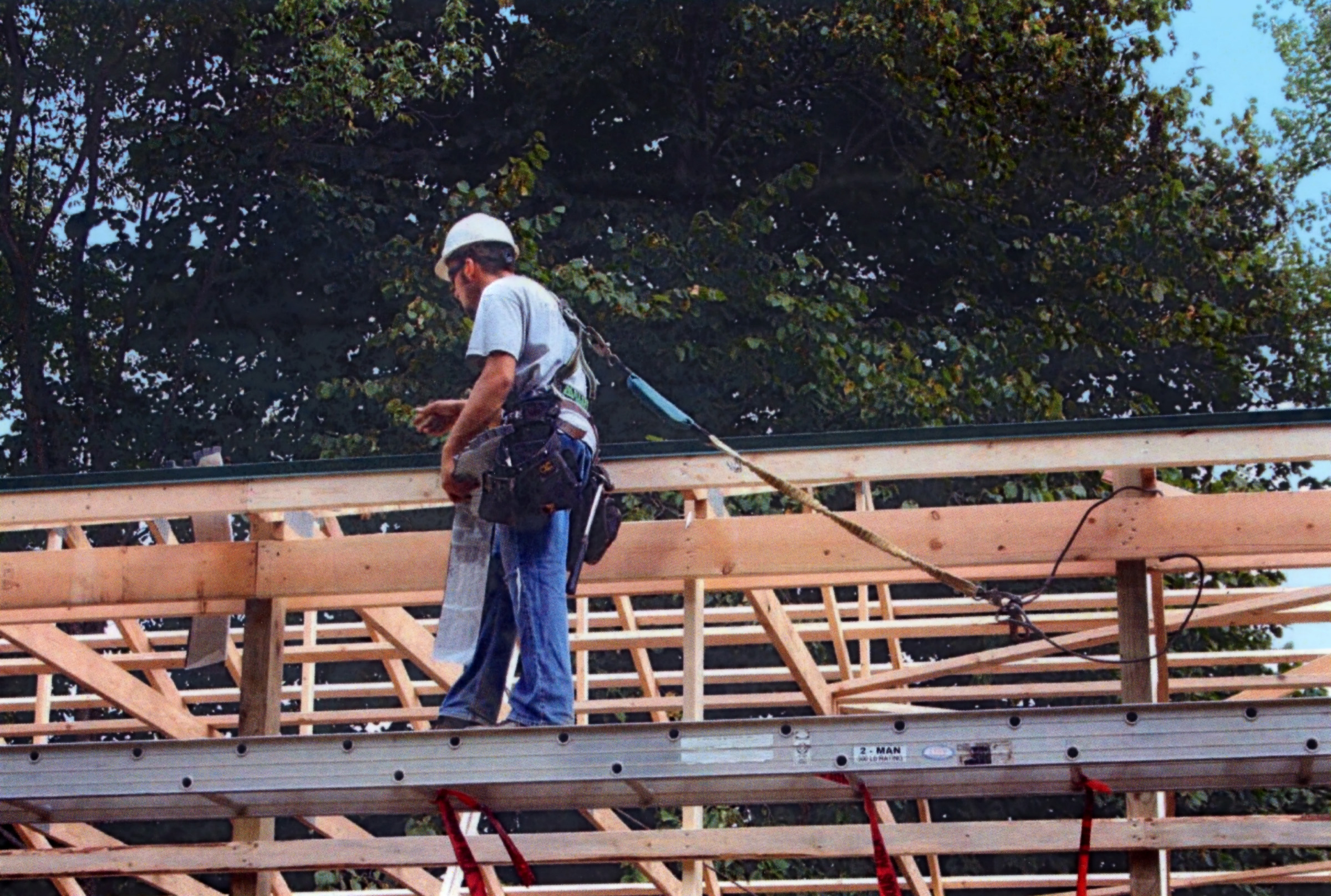 A worker at a residential construction site walks along a raised ladder placed horizontally while wearing a harness connected to an anchorage system.