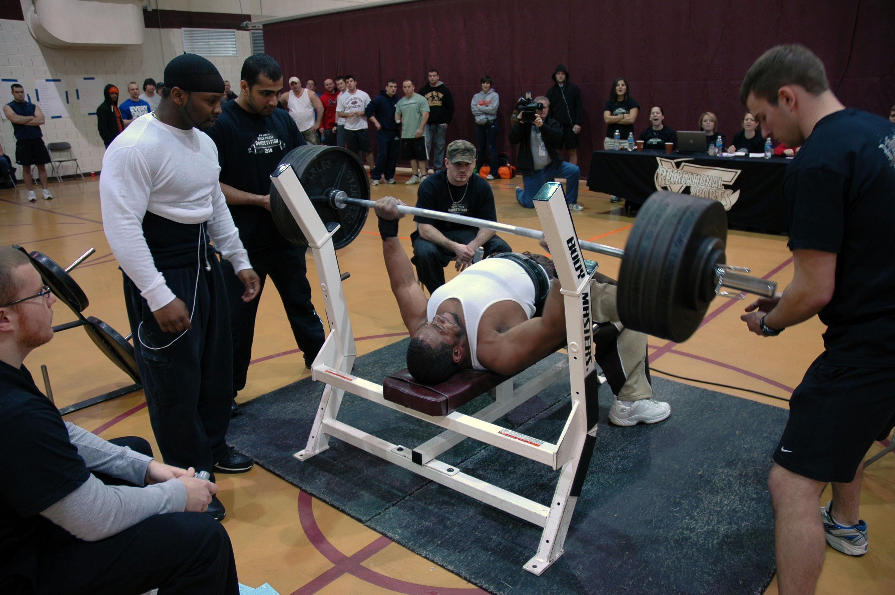 Photo of a competitor on the weight bench lifting a weight with a spotter on either side.