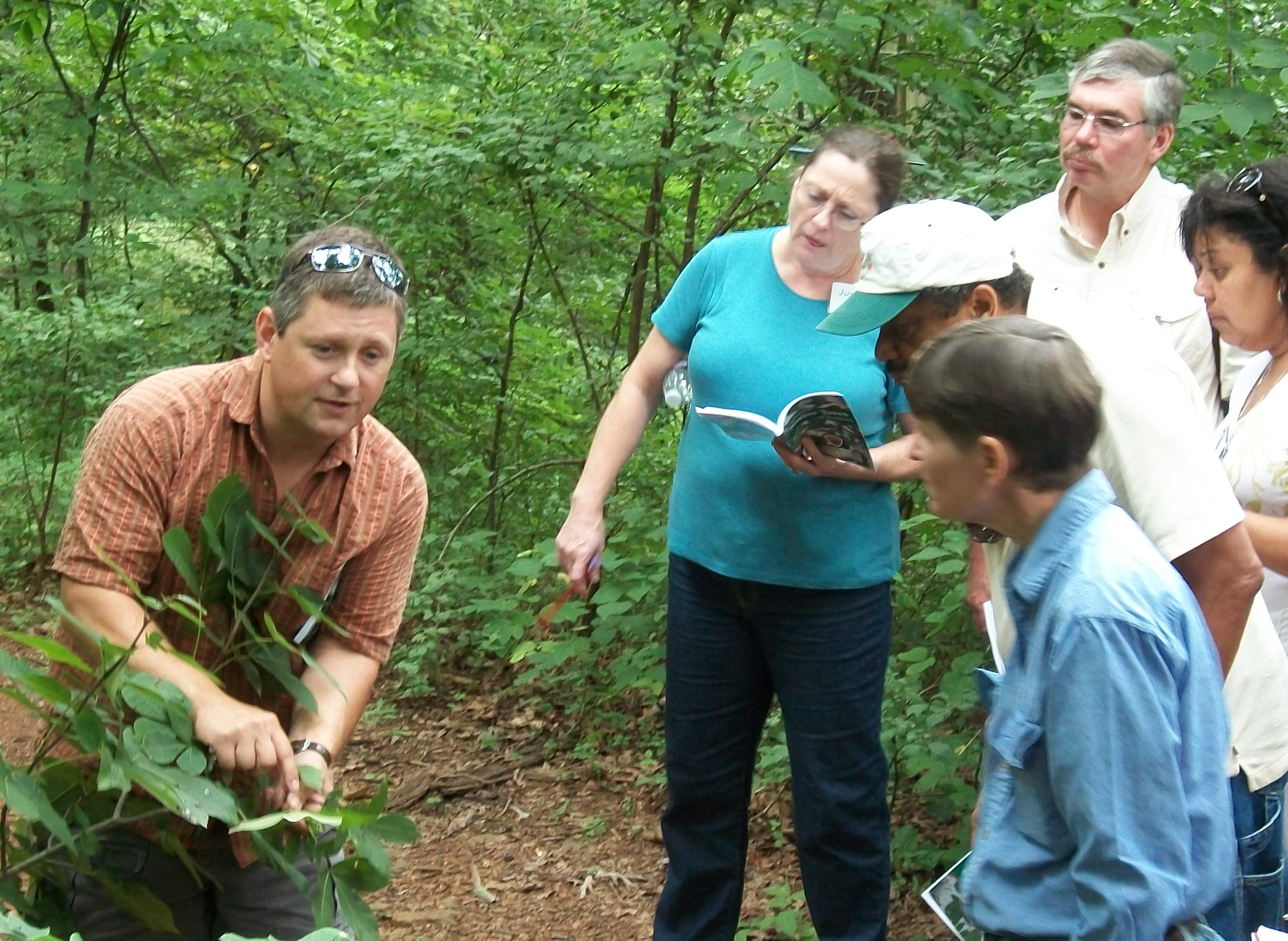 In a forest setting, John Peterson points to leaves on a bush while students look on.