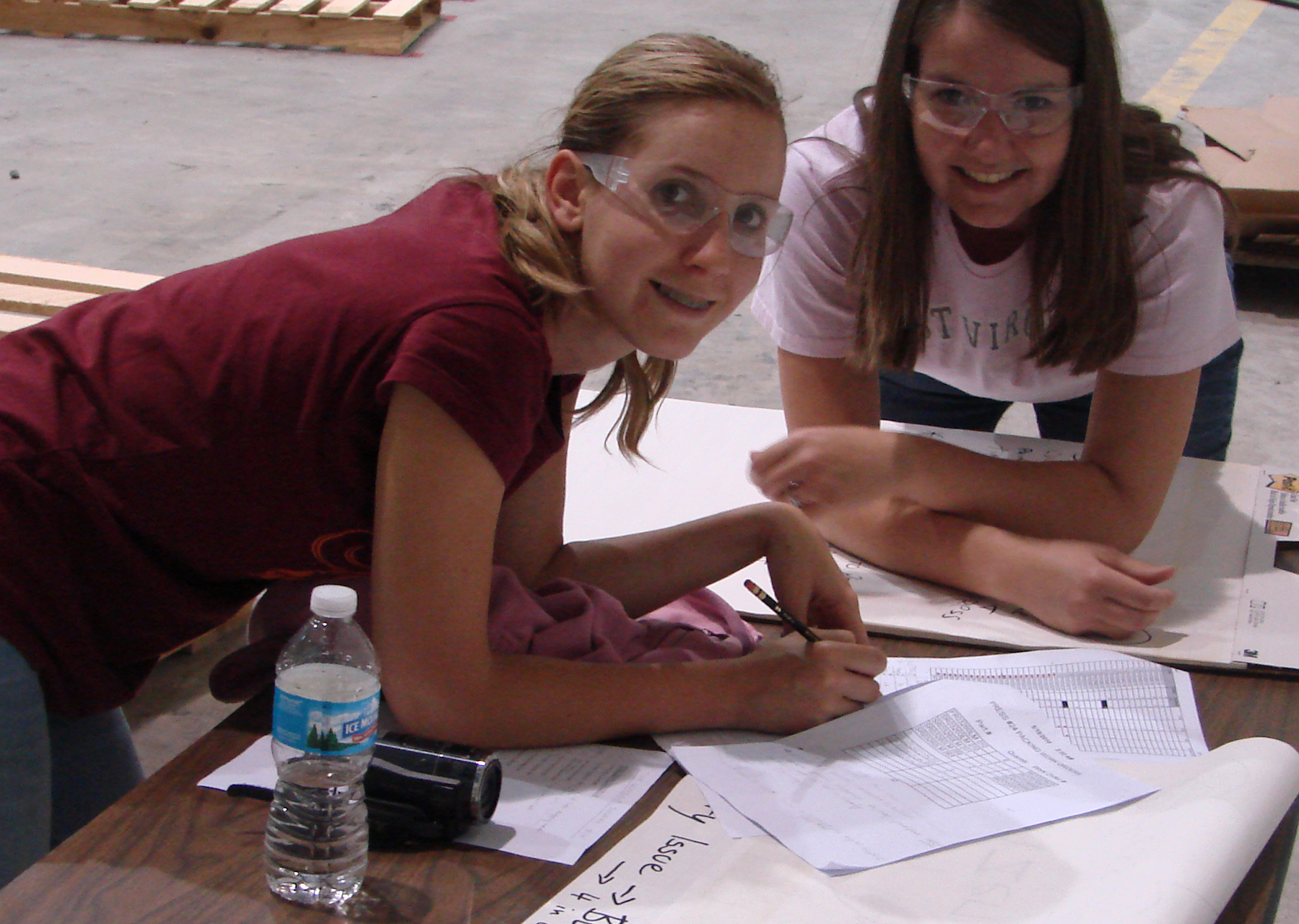 Two women wearing safety goggles write on papers covering a table on the Swan Corporation plant floor.