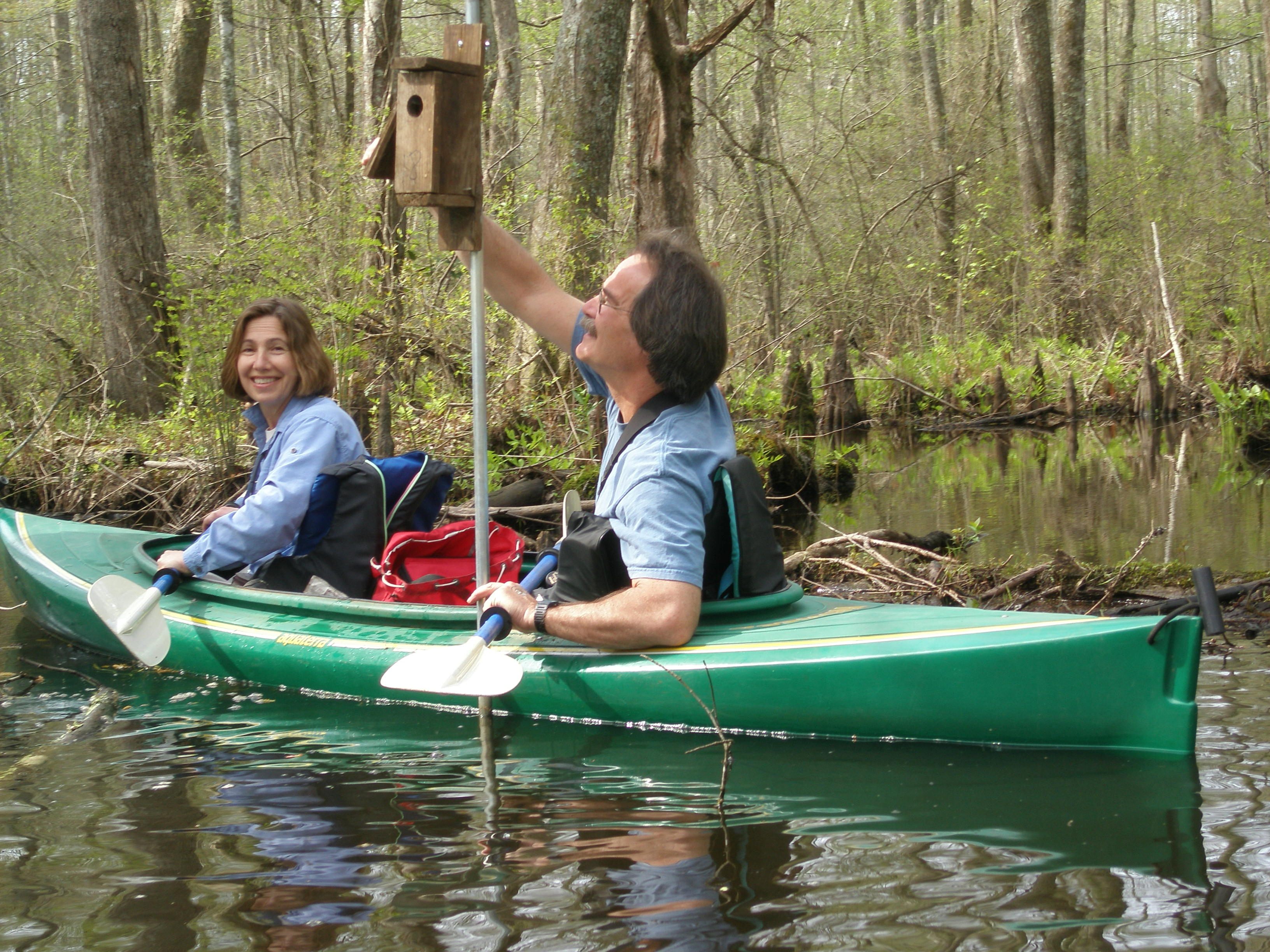 Two people in a kayak on a woodland pond, next to a bird box supported on a post.