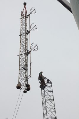 A worker is perched atop the lower section of a radio tower waiting to guide the upper section into place that is being lowered from above by a crane.