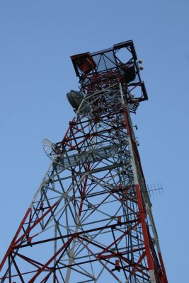 The view of a red and white radio tower from the ground up with a perfect blue sky in the background.