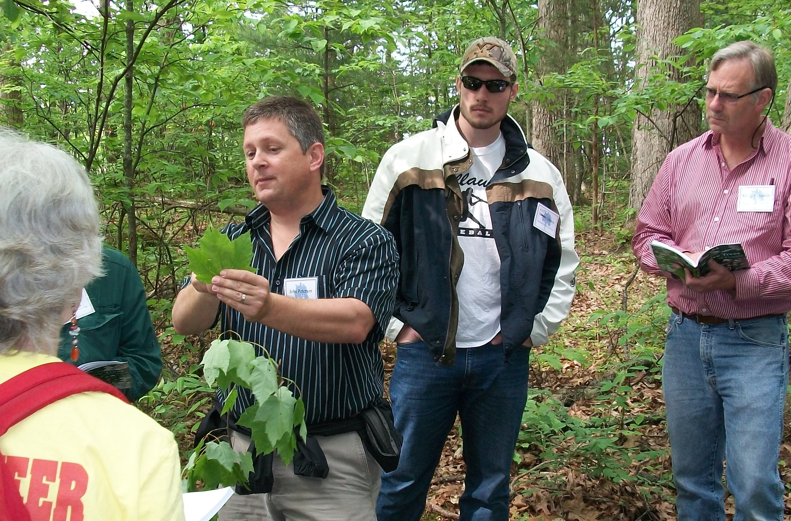 A group of people standing in a wooded setting; one is pointing a leaf in his hand.