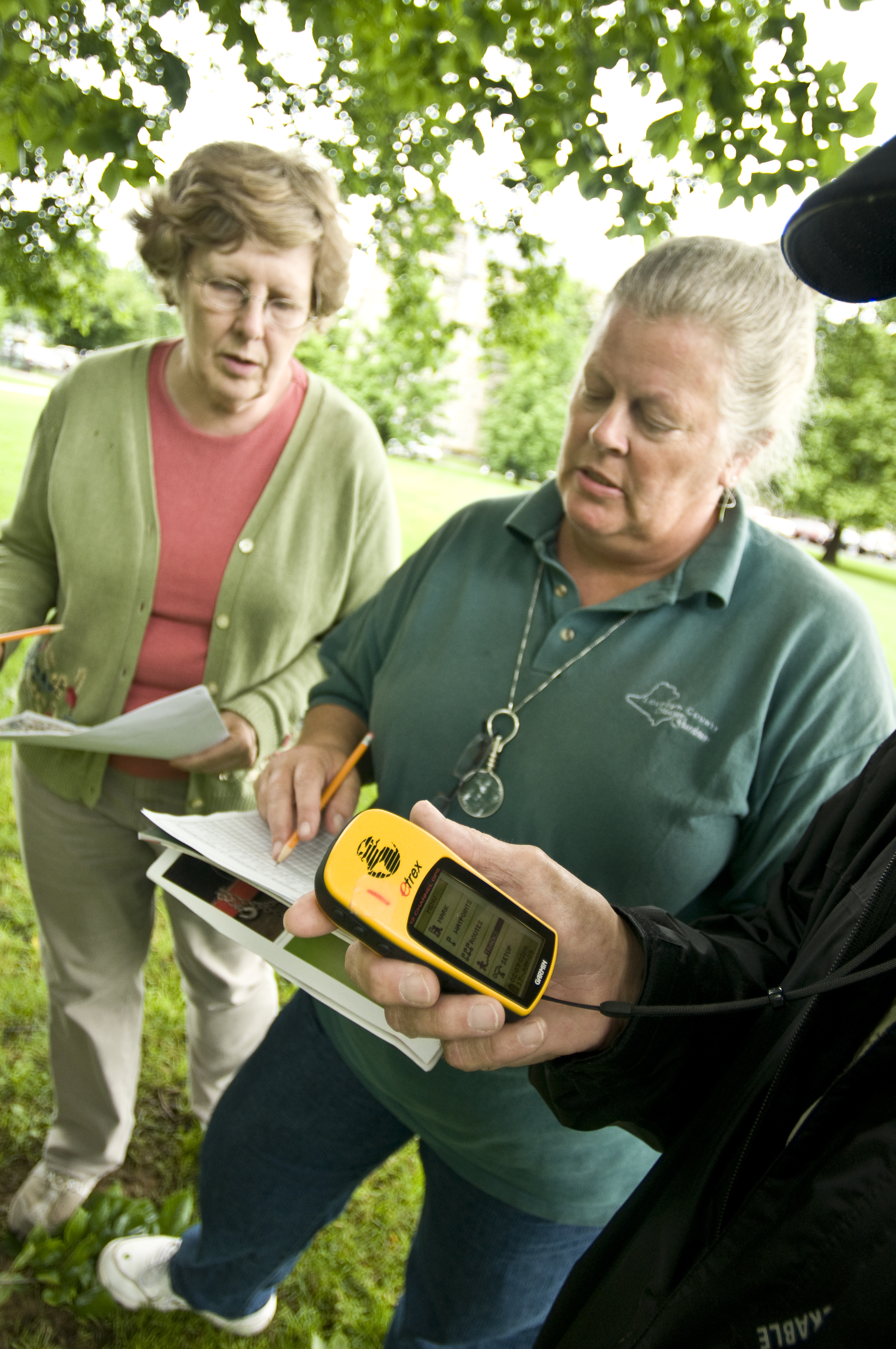Three people standing under a tree. Two hold papers and one holds a small GPS unit.