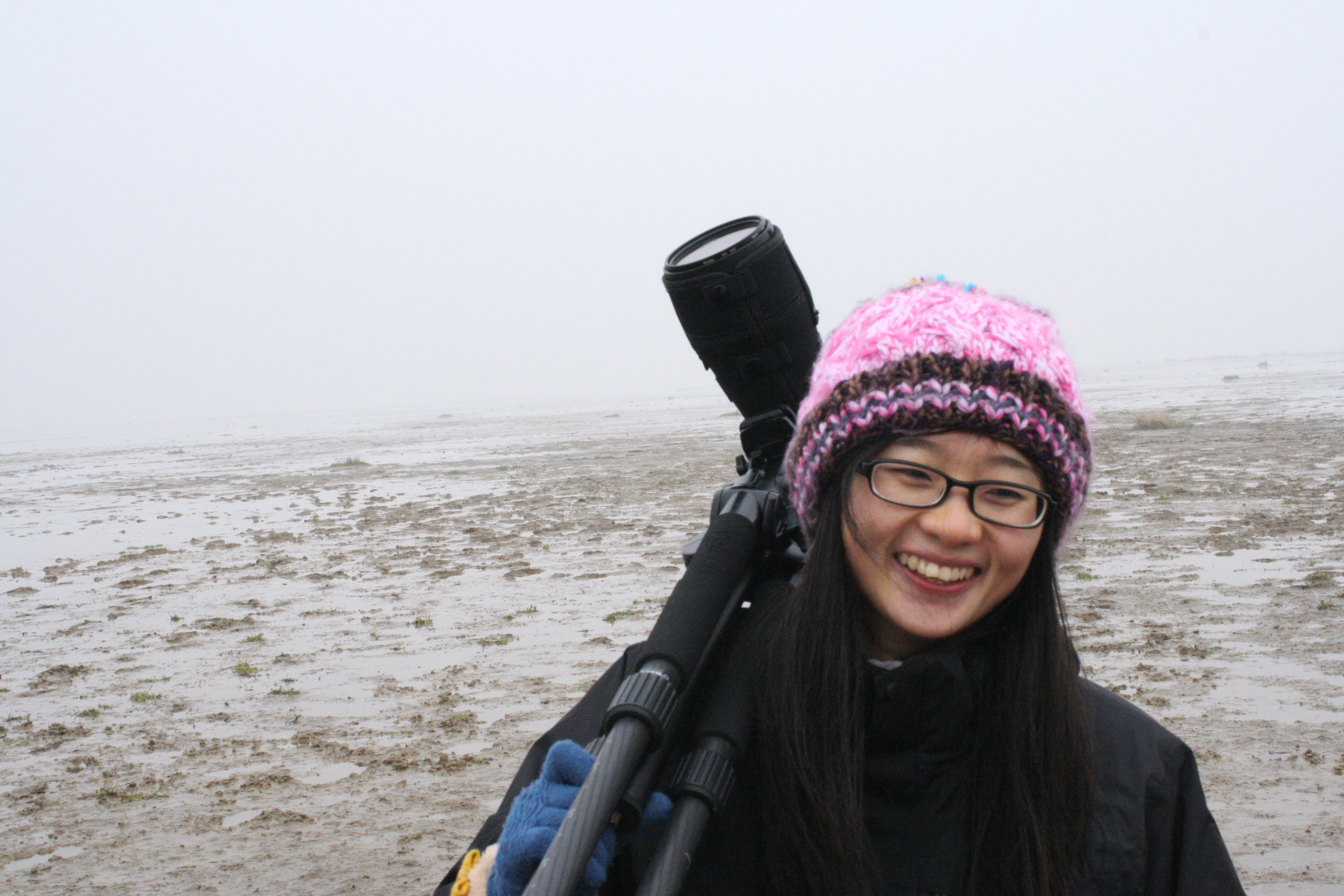 A woman holding a spotting scope stands on a mud flat