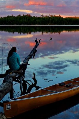 A lake at sunset with a girl and a canoe in the foreground.