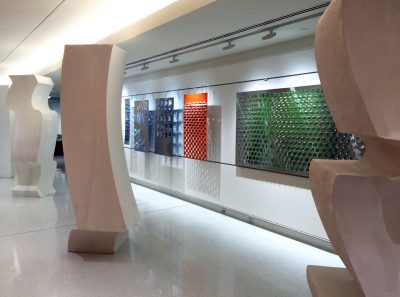 Large column sculptures in the foreground with colorful metal screens hung along a wall. 