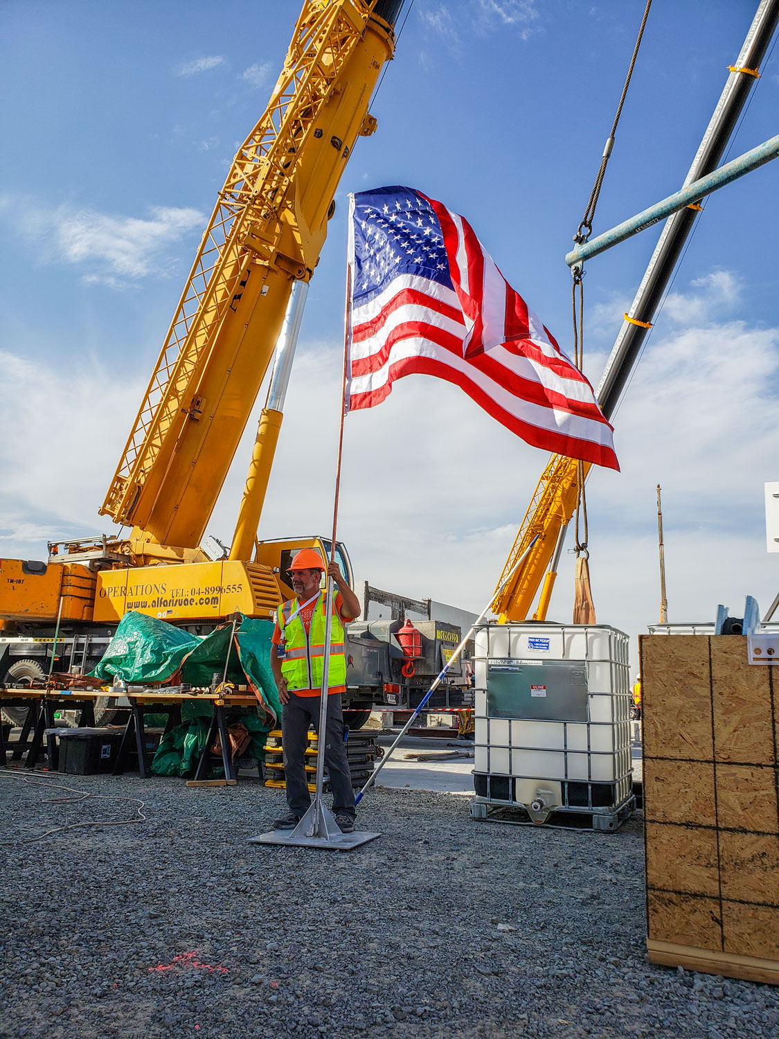 A man stands on gravel holding a flag pole with an American flag attached. Cranes stand behind him on an active construction site.