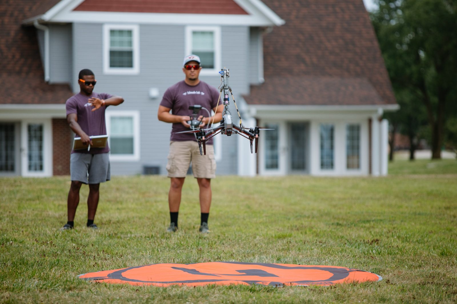 Undergraduate student Bryan Bloomfield (left) from Morehouse College examines his watch as David Schmale (right) in manning a sensor-mounted drone. An orange circle with an "H" is located underneath the hovering drone. Courtesy of Christina+David.