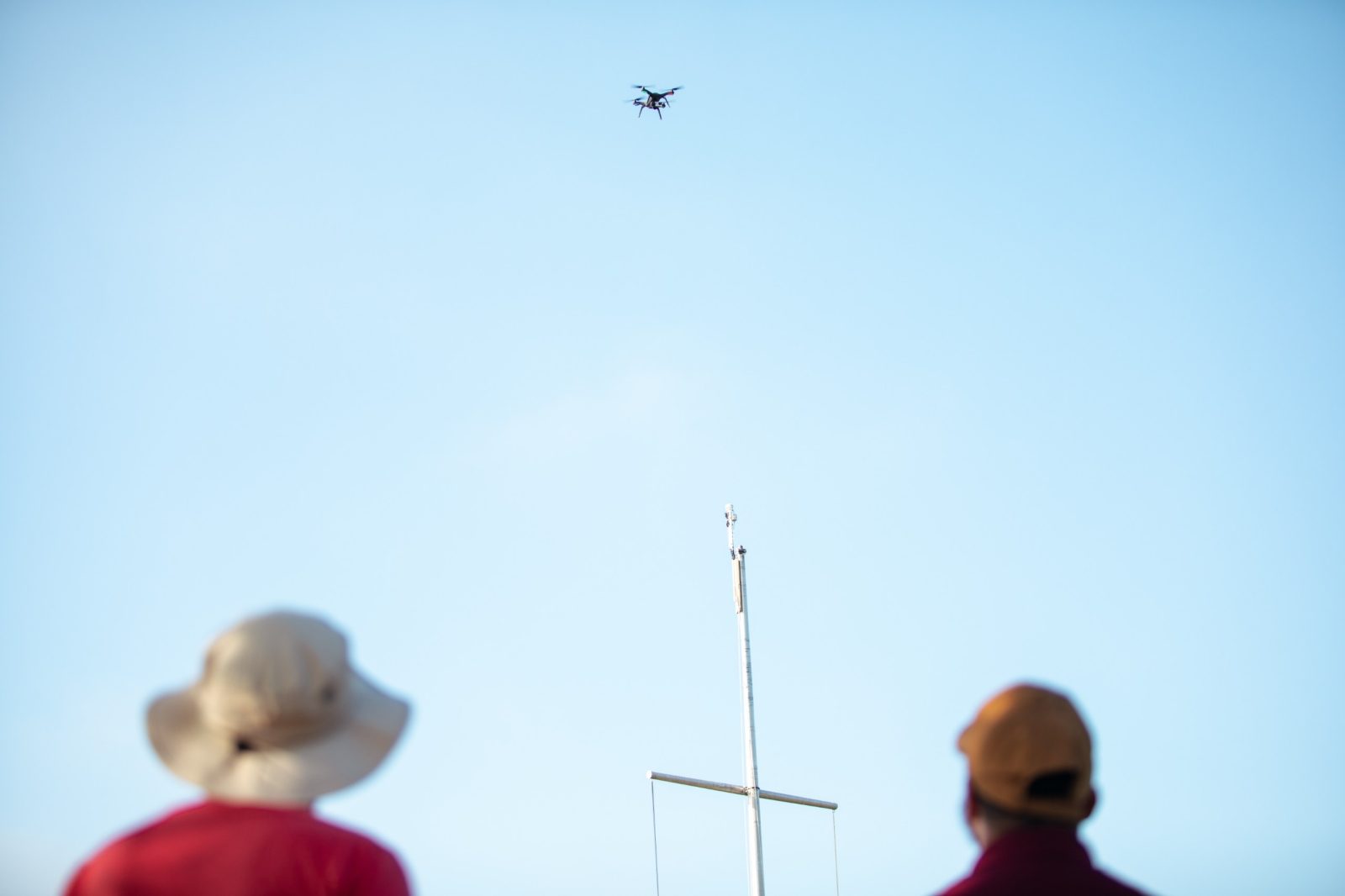 Virginia Tech graduate students Landon Bilyeu (left) and Javier Gonzalez-Rocha (right) use a drone to monitor wind at Grand Lake St. Marys, Ohio. A black drone hovers above a white nautical telescoping flagpole, with a brilliantly clear blue sky creating a stark contrast. Courtesy of Christina+David.