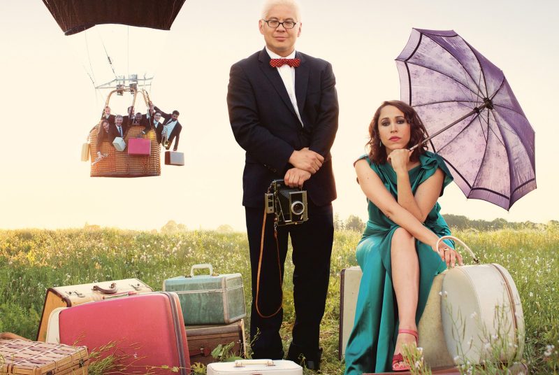 Posing in a field of grass and flowers in front of a hot air balloon and surrounded by luggage is Thomas Lauderdale holding a camera and China Forbes holding an umbrella.