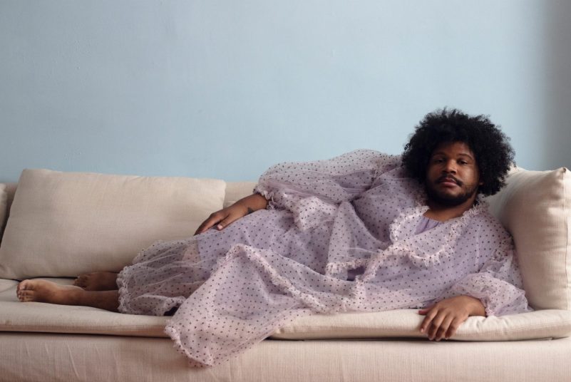A Black man wearing a sheer pink robe reclines on a beige sofa