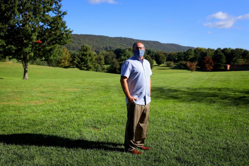 A man wearing a face covering stands in a large mowed field with trees and mountains in the distance