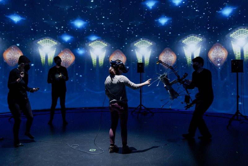 Researchers dressed in black hold various props as a user navigates the dark space decorated with colorful and unusual shapes.