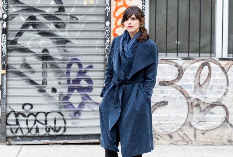 Author Valeria Luiselli stands outside in front of an industrial-looking wall covered in street art with a dark blue sashed overcoat and matching scarf.