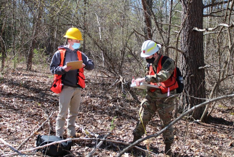 Two young men wearing hard hats, reflective vests, and face coverings and holding clipboards take notes in a wooded area.