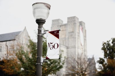 A banner celebrating Virginia Tech's 150th anniversary hangs on a lamp post with Burruss Hall in the background.