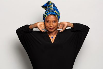 Singer Angelique Kidjo, dressed in a flowing black sweater, colorful head scarf, and a big blue gemstone around her neck, poses against a white wall with her hands touching the back of her neck.