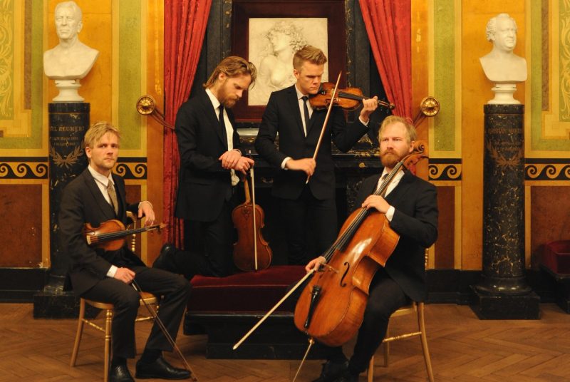 The members of the Danish String Quartet pose in a colorful museum-looking environment dressed in black suits. Two musicians sit in chairs, while two stand - all holding their instruments.,