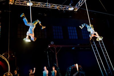 Two acrobats are suspended upside down by ropes in the air while other performers standing on the stage look up at them.