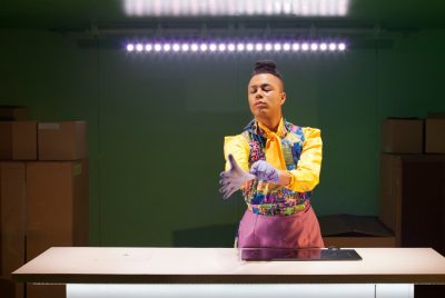 Performer Travis Alabanza wears a bright yellow shirt and multi-colored vest while snapping a purple rubber glove on his hand standing at a kitchen counter with a bright fluorescent light and stacks of boxes behind him.