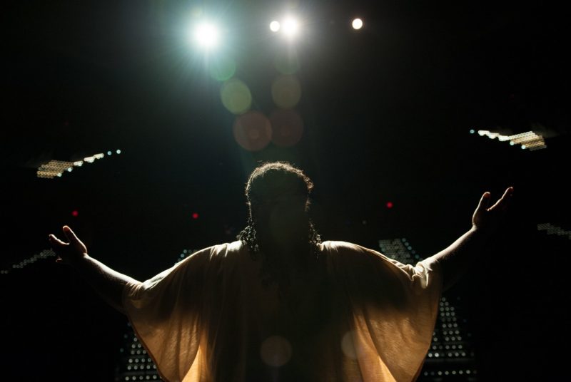 The back of a person dressed in a sheer, white, flowing top stretches their hands out and up in the air while standing on a dark stage, bright lights shine above their head.