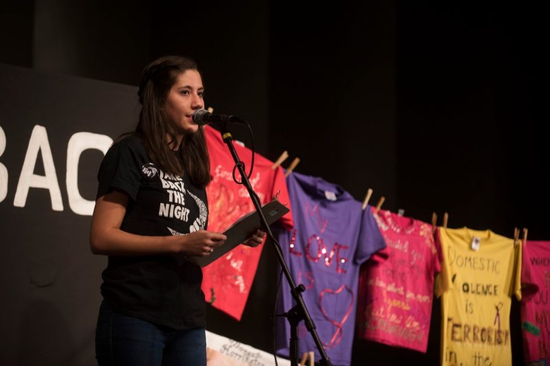 A student speaks at a microphone at a Take Back the Night Rally, with t-shirts from the Clothesline Project hanging behind her.