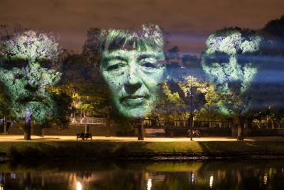 Faces of three individuals are projected onto the foliage of three trees facing a stream.