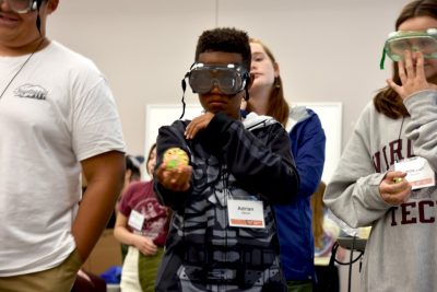 A participant in the middle-school-focused Explore Science tries to aim and throw a small plastic ball into a wastebasket while wearing a pair of special plastic googles that skew his vision. 