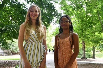  Janey Dike and Brianna George, both wearing summer dresses, stand behind Williams Hall, with trees covering the background.