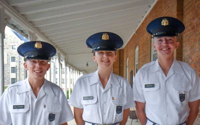 Three smiling cadets stand shoulder to shoulder in white unform shirts and blue military hats on the covered porch of Lane Hall. Pearson Hall West is in the background.