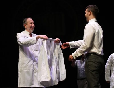 VTCSOM Dean Lee Learman holds out a new white coat to the outstretched hand of a first-year student.