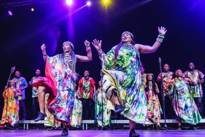 Two members of Soweto Gospel Choir dance at the front of a stage. They both have one foot off the ground and their hands in the air. They're wearing one-shouldered, flowing robes with beaded jewelry on their wrists and around their necks. Behind them, the rest of the choir sings in colorful attire.