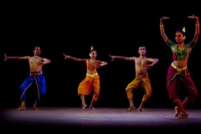 Performers with two dance ensembles perform onstage. A woman stands in front, wearing silky, flowing maroon pants and a silky green cropped top, gold jewelry on her wrists and neck. With her feet together, but her legs bent she reaches above her head with her hands. Behind her, three dancers move. One on the far left is a shirtless man with silky electric blue pants, the woman in the middle wears flowy orange pants with a matching cropped top, and a second shirtless man dances in silky yellow pants.