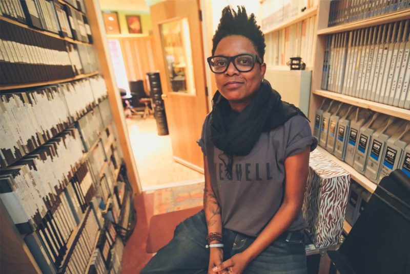 Artist Shirlette Ammons sits in a studio, surrounded by shelves filled with albums and cartridges. She's wearing a gray t-shirt, jeans, a black scarf, and dark-rimmed glasses.