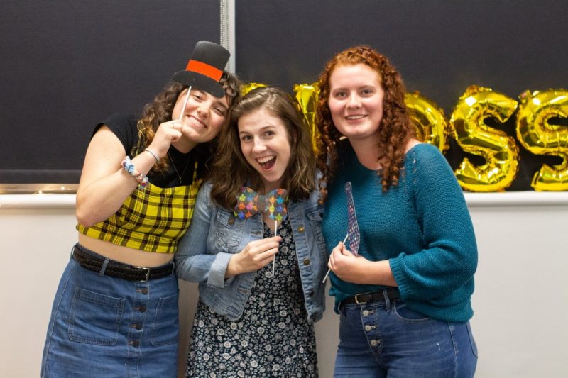 Three Virginia Tech undergraduates hold up photo booth props (paper hats and bow ties on sticks) in front of a chalkboard at Virginia Tech's Glossolalia Festival.