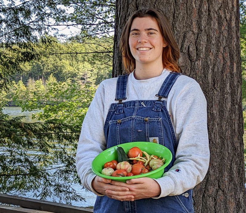 Lou Groundwater, in denim overalls, holds a bowl of vegetables at camp.