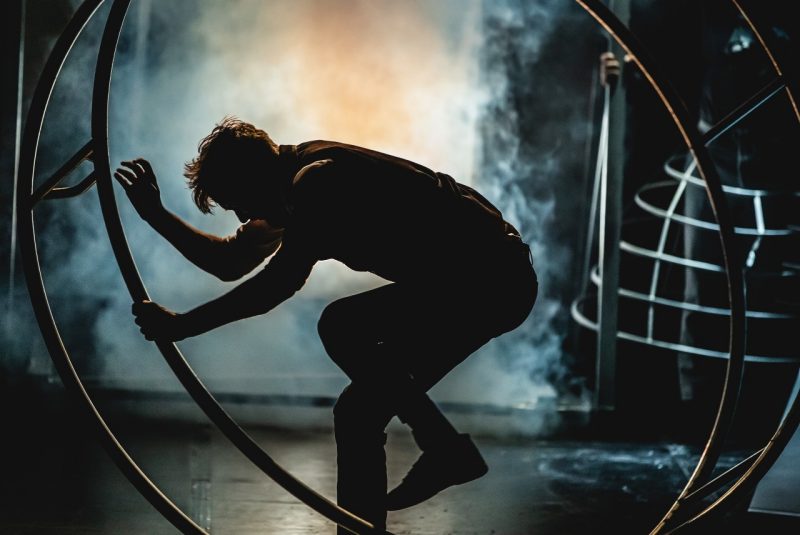 A silhoutted cirque performer steps into a circular wheel, preparing for a stunt.