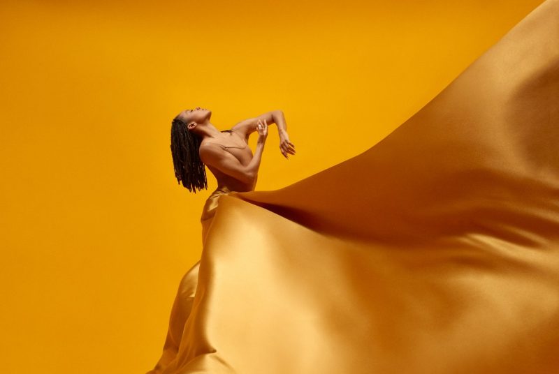 An Alvin Ailey dancer poses with her head up to the sky, eyes closed, long hair falling down her back, arms out in front of her like she's embracing the air. She wears a gold satin dress with a huge skirt that flies up in the air in front of her. The background is a rich gold.
