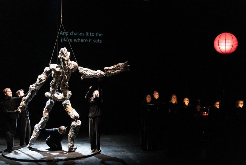 Puppeteers perform "Book of Mountains and Seas" on stage. On the left, lit with white light from above, puppeteers maneuver a large puppet that somewhat resembles diftwood and somewhat resembles a human figure using ropes on pulleys. On the right is a red round paper lantern. above a dimly lit chorus of singers.